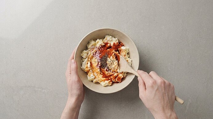 Zwei Hände rühren mit einem Holzlöffel in einer Schüssel weißer, krümeliger Masse, bedeckt mit roter Sauce und Gewürzen.