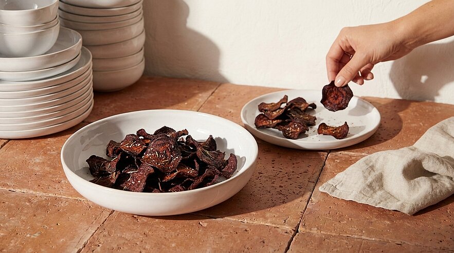 Zwei weiße Schalen mit dunklen, knusprigen Gemüsescheiben (Chips); eine Hand nimmt eine Chips heraus.