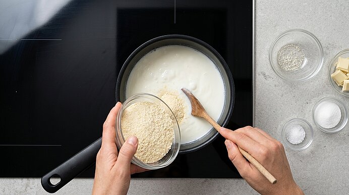 Draufsicht: Hände rieseln Riebelmais aus einer Schale in heiße Milch im Topf und rühren mit einem Holzlöffel.