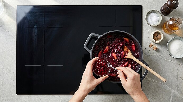 Hände geben Cranberries in einen Topf mit köchelnder roter Sauce auf einem schwarzen Kochfeld, ein Holzlöffel rührt darin.