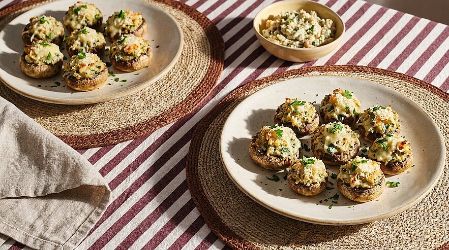 Zwei Teller mit gebackenen, mit Kräuter‑Käse‑Füllung gefüllten Champignons, bestreut mit geriebenem Käse und gehackter Petersilie.