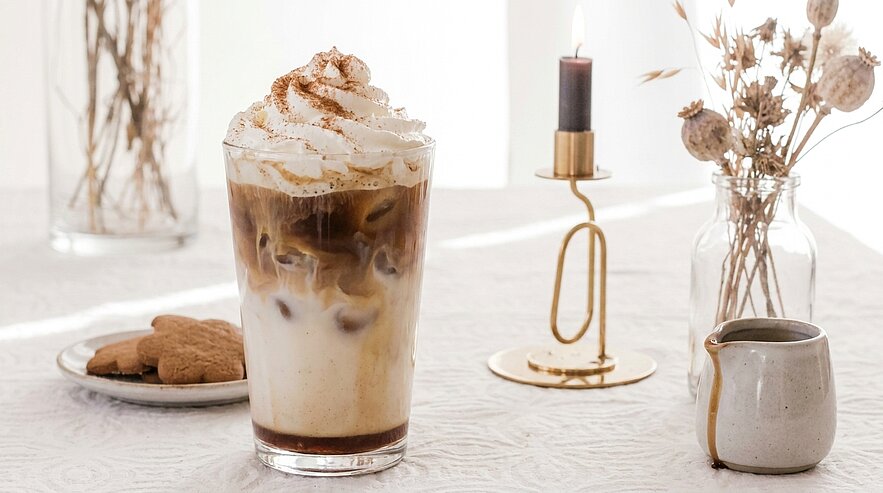 Glas Eiskaffee mit geschichteten Milch- und Kaffeeschichten, Eiswürfeln, Sahnehaube und Zimtpulver; daneben Teller mit Keksen und kleines Milchkännchen auf einem Tisch.