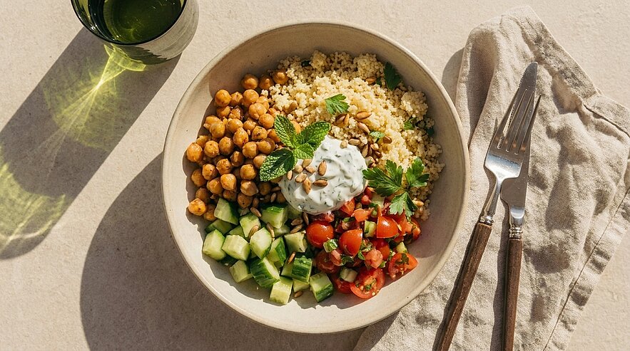Schale mit Couscous-Bowl: gebratene Kichererbsen, gewürfelte Gurke, halbierte Kirschtomaten, Kräuter-Joghurt, frische Kräuter und Pinienkerne.