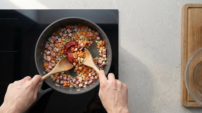 Zwei Hände rühren mit Holzlöffeln in einer Pfanne gewürfelte Karotten, Zwiebeln und Schinken; Tomatenmark in der Mitte.