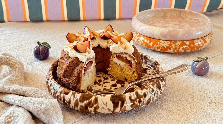 Ein frisch gebackener Kuchen mit einer hellen Oberseite, dekoriert mit Sahne und Scheiben von Früchten. Ein Stück des Kuchens wurde herausgeschnitten und auf einem dekorativen Teller platziert. Im Hintergrund sind bunte Streifen und ein weiterer Teller zu sehen.