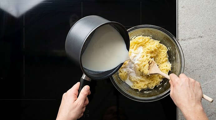 Hände gießen heiße Milch aus einem kleinen Topf in eine Glasschüssel mit gestampften Kartoffeln und rühren die Mischung mit einem Holzlöffel.