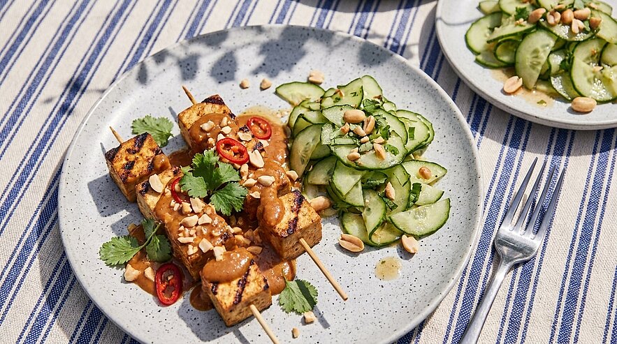 Teller mit drei gegrillten Tofu-Spießen, überzogen mit Erdnusssauce und garniert mit gehackten Erdnüssen, Koriander und roten Chilischeiben; dazu dünn geschnittener Gurkensalat mit Erdnussstücken.
