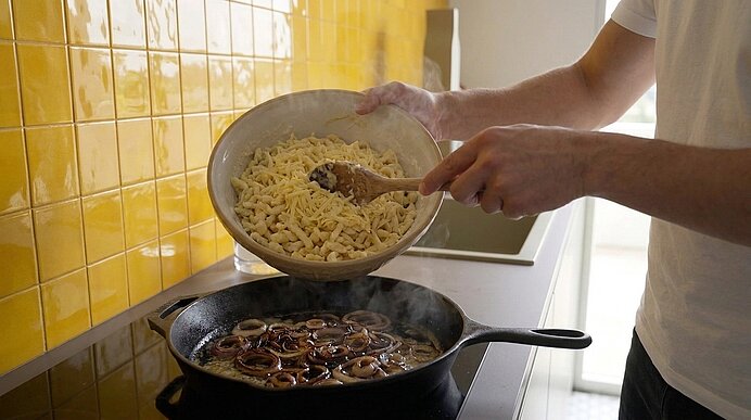 Hände einer Person kippen eine Schüssel mit geraspelter, heller Masse in eine Pfanne, in der Zwiebelringe goldbraun braten.