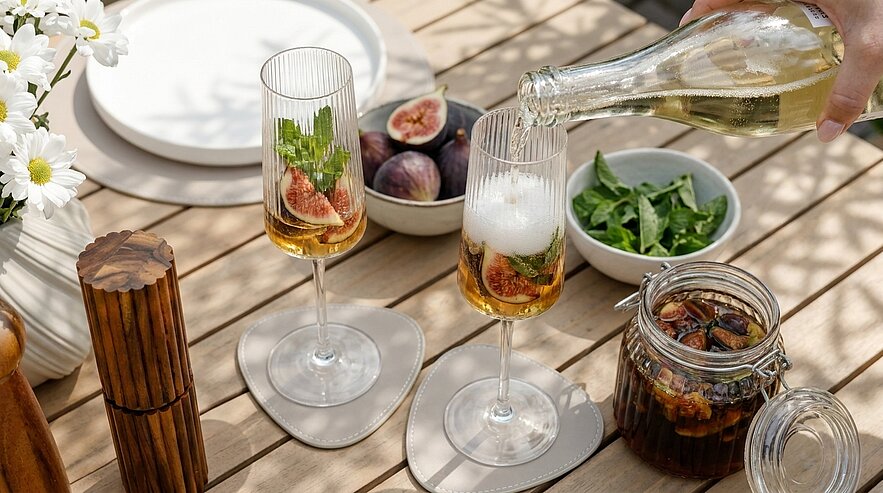 Hand gießt Sekt in zwei Gläser mit frischen Feigen und Kräutern auf einem Holztisch; daneben Schale mit Feigen, eine Schüssel Salat und ein Glas mit eingelegten Früchten.