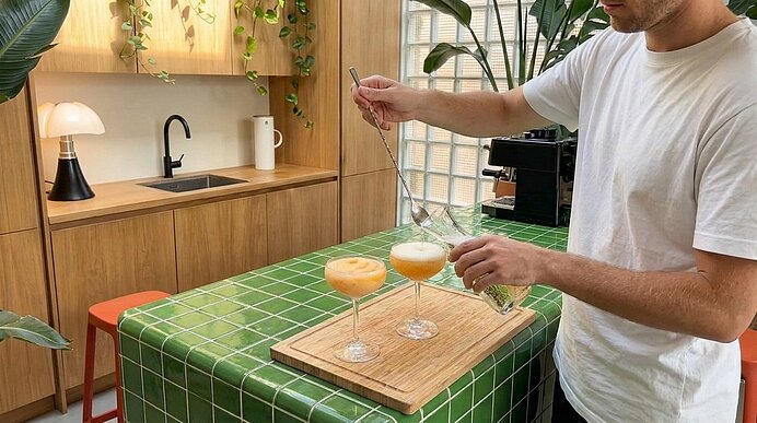 Person gießt aus einer Glas-Karaffe grünes Getränk in zwei Cocktails mit dünner Schaumkrone, die auf einem Holzbrett auf einem grün gefliesten Küchenblock stehen.