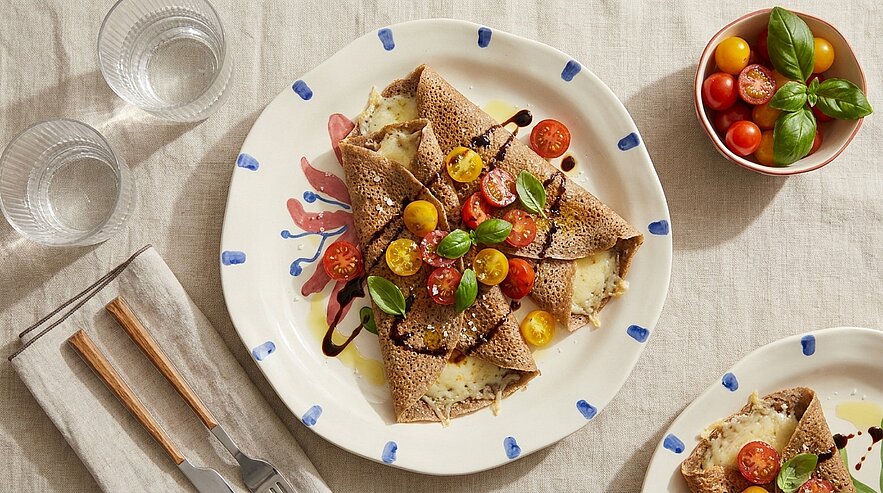 Drei herzhafte Buchweizen-Galettes (gefüllte Crêpes) auf einem Teller, mit geschmolzenem Käse, halbierten Kirschtomaten, frischem Basilikum und Balsamico-Reduktion garniert.