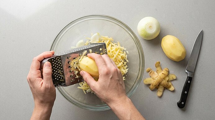 Zwei Hände reiben eine geschälte Kartoffel auf einer Reibe über einer Glasschüssel mit geriebenen Kartoffeln; daneben liegen eine Zwiebel, eine geschälte Kartoffel und ein Küchenmesser.