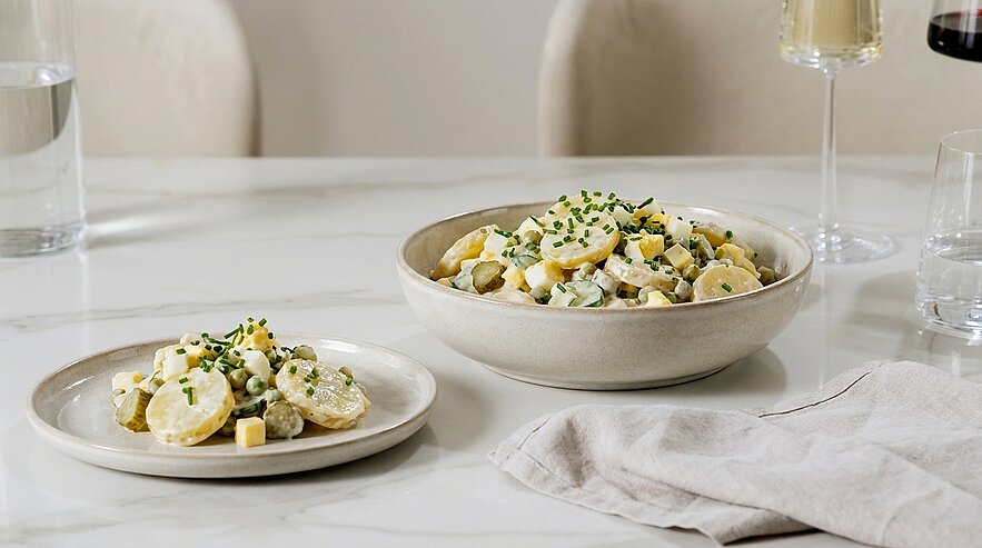 Schüssel und kleiner Teller mit cremigem Kartoffelsalat, garniert mit gehacktem Ei und Schnittlauch, auf hellem Tisch.