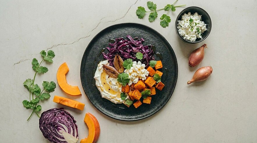Teller mit herbstlicher Bowl: gebackene Kürbiswürfel, geschmorter Rotkohl, cremiges Joghurtdressing und Hüttenkäse, garniert mit frischem Koriander.