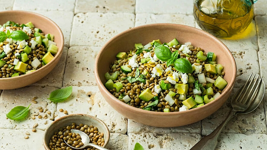 Eine Schüssel mit Linsensalat, angereichert mit grünen Äpfeln, Gurken und frischem Basilikum. Kleine Feta-Würfel ergänzen das Gericht. Im Hintergrund befindet sich ein Behälter mit Olivenöl und eine kleine Schale mit weiteren Linsen. Der Untergrund ist aus hellen Steinen.