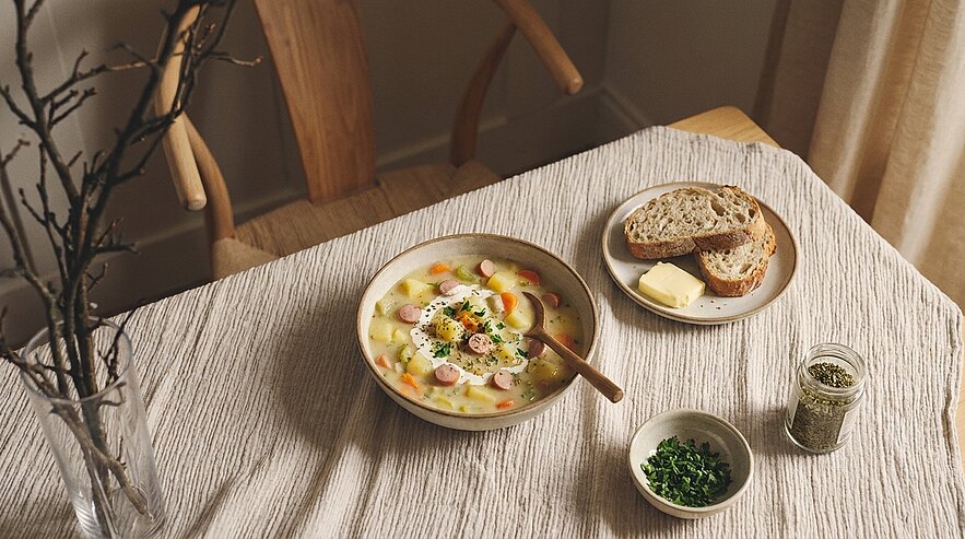 Gedeckter Tisch mit einer Schüssel cremiger Gemüsesuppe mit Wurst- und Eierscheiben, dazu Teller mit Brot und Butter sowie kleine Schälchen mit frischen und getrockneten Kräutern.