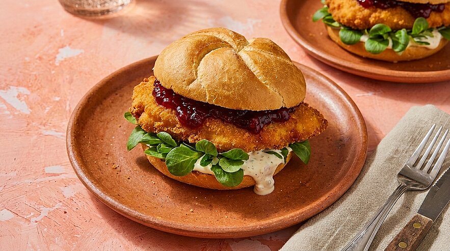 Brötchen mit knusprig paniertem Hähnchenfilet, frischen grünen Blättern, cremiger Sauce und dunkler Beerenglasur auf braunem Teller.