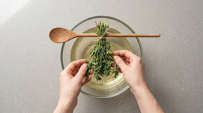 Zwei Hände halten Waldmeister kopfüber an einem Holzlöffel in eine Schüssel mit Wein auf hellgrauer Arbeitsplatte.