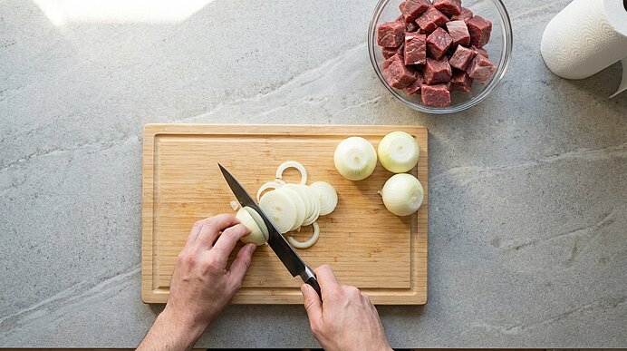 Hände schneiden eine Zwiebel in Scheiben auf einem Holzschneidebrett; daneben eine Schüssel mit gewürfeltem rohem Fleisch.