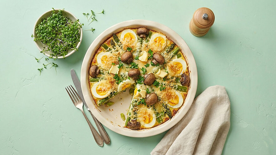 Ein Teller mit einer fleischlosen Quiche, belegt mit Spargel, Eiern, Champignons und Käse. Die Quiche hat eine goldbraune Kruste und ist in Stücke geschnitten. Neben dem Teller liegt eine Gabel und ein Messer auf einem hellen Servietten. Im Hintergrund ein Schälchen mit frischen Kräutern.
