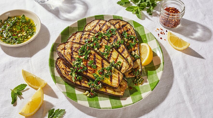 Gegrillte Auberginenscheiben auf einem grün-weiß karierten Teller, garniert mit gehackten Kräutern, Chiliflocken und Olivenöl, dazu Zitronenspalten und ein Kräuter-Dip.