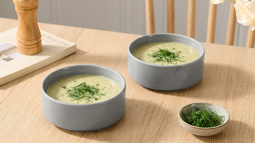 Zwei graue Schalen mit cremiger Suppe, garniert mit frischem Dill, stehen auf einem Holztisch. Daneben befindet sich eine kleine Schale mit zusätzlichem Dill. Der Hintergrund ist hell und schlicht.