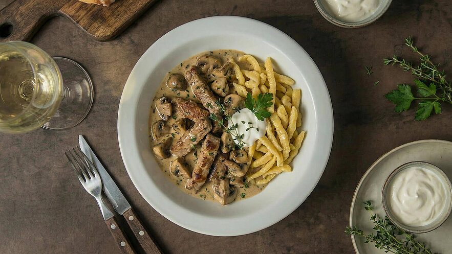 Ein Teller mit saftigen Fleischstreifen in einer cremigen Pilzsauce, serviert mit frischen Spätzle. Ein Klecks saure Sahne und ein paar Blätter Petersilie garnieren das Gericht. Daneben ein Glas Weißwein und einige Kräuter. Der Tisch hat einen rustikalen Holzlook.