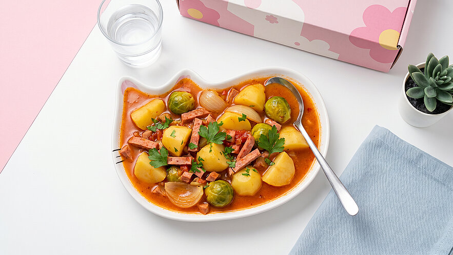Ein Teller mit einer bunten Mischung aus Kartoffeln, Würstchen, Rosenkohl und Zwiebeln in einer würzigen Sauce. Das Essen ist mit frischer Petersilie garniert. Daneben steht ein Glas Wasser und ein kleiner Topf mit einer Pflanze.