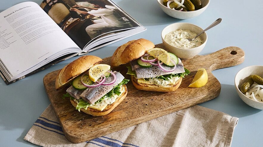 Zwei Fischbrötchen auf einem Holzbrett: Brötchen mit Salat, Heringsfilets, Gurkenscheiben, roten Zwiebeln und Zitronenscheiben; daneben Schale mit Remoulade und eingelegten Gurken.