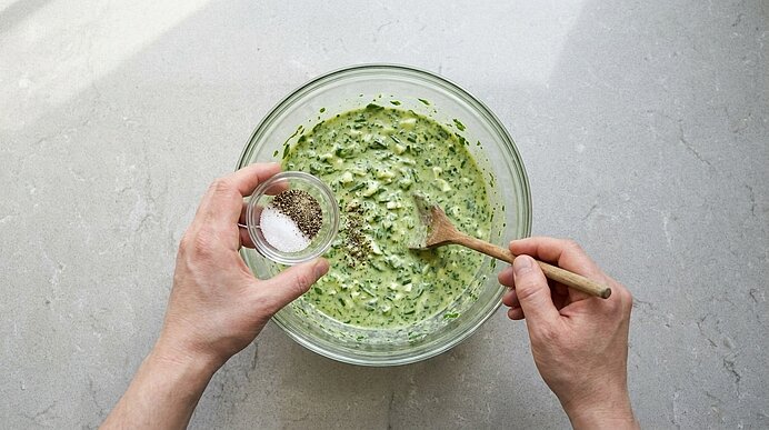 Zwei Hände über einer Glasschüssel mit grünem Kräuterdip; eine Hand hält ein kleines Schälchen mit Salz und Pfeffer, die andere rührt mit einem Holzlöffel.
