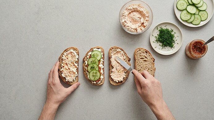 Zwei Hände bestreichen Brotscheiben mit Frischkäsemischung und belegen sie mit Gurkenscheiben und Dill.