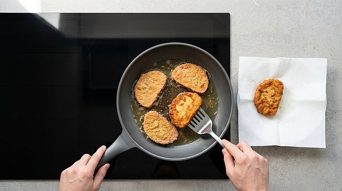 Hände wenden panierte, goldbraun gebratene Schnitzel in einer Pfanne auf dem Herd; vier Stücke braten, eines liegt auf Küchenpapier zum Abtropfen.