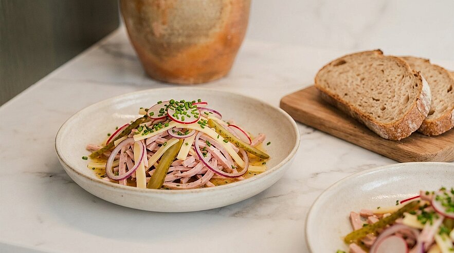 Teller mit Wurstsalat aus dünnen Wurststreifen, Gewürzgurken, roten Zwiebeln, Schnittlauch und Öl-Dressing; daneben mehrere Brotscheiben.