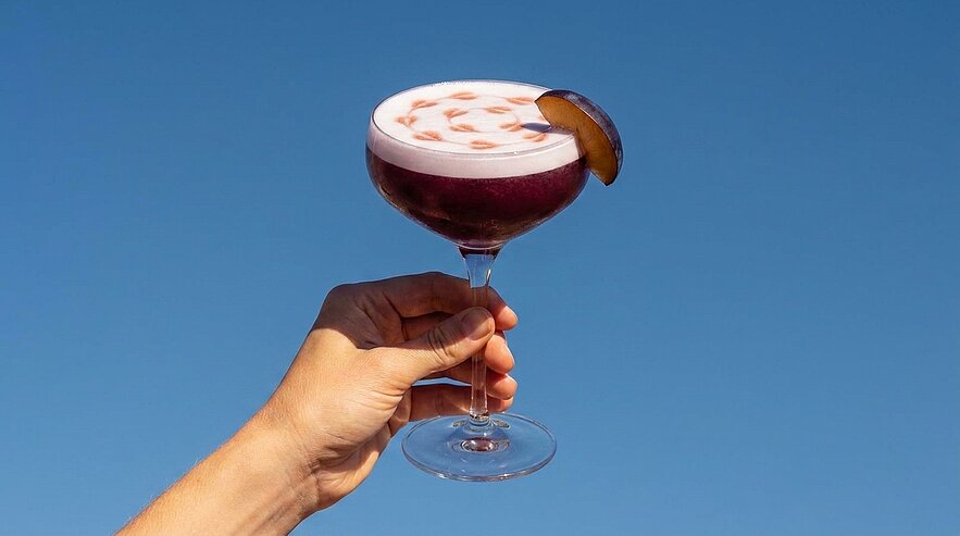 Hand hält eine Coupette mit dunkelviolettem Cocktail, schaumiger weißer Schicht mit Bitterspritzmuster und einer Orangenscheibe als Garnitur vor blauem Himmel.