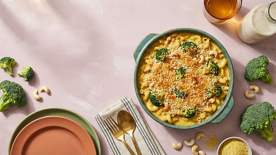 Ein Auflaufgericht in einer blauen Schüssel, überbacken mit einer goldenen Kruste, und verziert mit Brokkoli und Cashewkernen. Daneben liegen zwei Teller, Besteck und eine Streuung von Nahrungsmitteln wie Brokkoli und Cashewkerne auf einer hellen Oberfläche.
