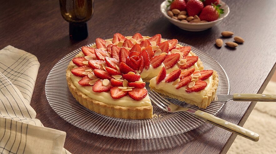 Fruchtige Erdbeertarte auf Glasplatte, ein Stück entnommen und auf Tortenschaufel, cremige Füllung und knuspriger Mürbeteig, oben reichlich mit frischen Erdbeerscheiben dekoriert.