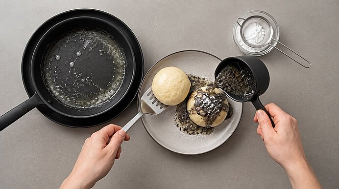 Zwei Hände träufeln flüssige Mohnbutter aus einem kleinen Topf über gegarte Germknödel in einer Pfanne.