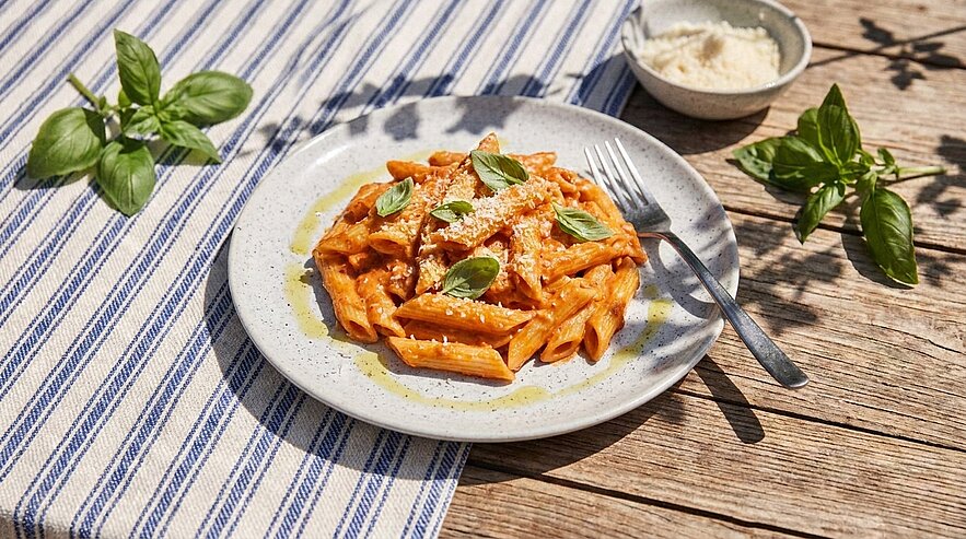 Teller mit Penne in cremiger Tomatensauce, bestreut mit geriebenem Parmesan und frischen Basilikumblättern; eine Gabel liegt daneben.