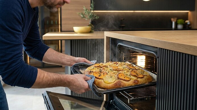 Person zieht eine frisch gebackene, mit Kartoffelscheiben und Kräutern belegte Focaccia aus dem heißen Ofen, Dampf steigt auf.