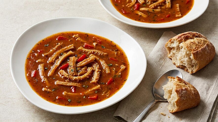 Teller mit Kuttelsuppe: Streifen von Kutteln in würziger, tomatenfarbener Brühe mit roten Paprika und Kräutern, daneben ein zerbrochenes knuspriges Brötchen und Löffel.