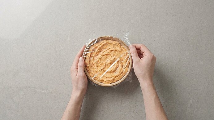 Zwei Hände ziehen Frischhaltefolie über eine runde Tarte mit cremiger Füllung auf einer hellen Arbeitsfläche.