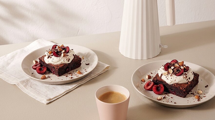 Zwei Teller mit Schokoladenbrownies, jeweils mit Schlagsahne, halbierten Kirschen und gehackten Nüssen garniert; daneben eine Tasse Kaffee.
