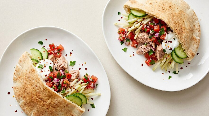 Zwei weiße Teller mit gefüllten Pitataschen: Thunfisch, gewürfelte Tomaten, Gurkenscheiben, feine Salatstreifen und Joghurt-/Tzatziki-Dip, garniert mit Kräutern und Chiliflocken.