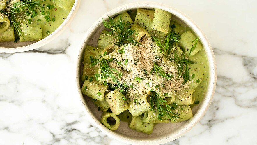 Eine Schüssel mit Rigatoni-Pasta in einer cremigen grünen Sauce, garniert mit frischem Dill und gehacktem Schnittlauch. Die Oberfläche ist mit zerstoßenen Nüssen bestreut, was eine zusätzliche Textur verleiht. Der Hintergrund ist eine helle, marmorierte Oberfläche.
