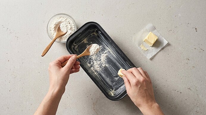 Zwei Hände fetten eine Kastenform: eine reibt Butter ein, die andere streut Mehl aus einem Löffel; daneben liegt ein Butterstück auf Papier und eine Schüssel mit Mehl.
