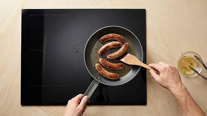 Zwei Hände braten vier goldbraune Bratwürste in einer Pfanne auf einem schwarzen Kochfeld; ein Holzspatel wendet eine Wurst.