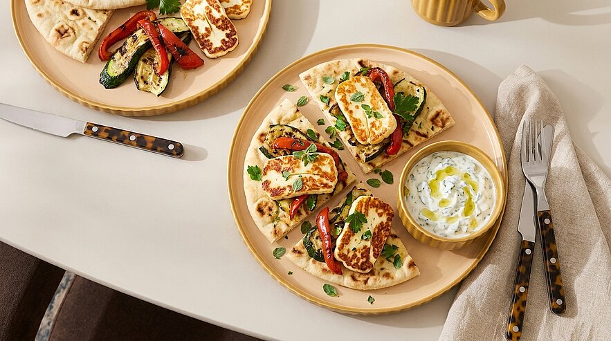 Teller mit Fladenbrot belegt mit gegrilltem Halloumi, gebratenem Gemüse (Zucchini, Paprika) und frischer Petersilie, dazu ein Schälchen Joghurt-Dip mit Olivenöl.