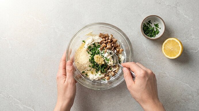 Zwei Hände verrühren Frischkäse, Parmesan, Kräuter und Champignonstiele in einer Glasschüssel mit einem Löffel.