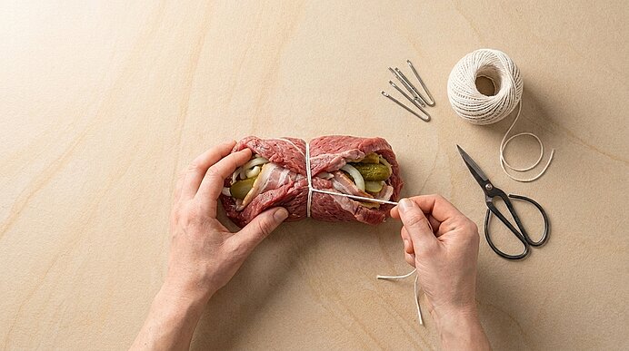 Hände binden mit Küchengarn eine gefüllte Rinderroulade (mit Gurken und Zwiebeln) auf einem hellen Holzbrett; Schere und Garn liegen daneben.