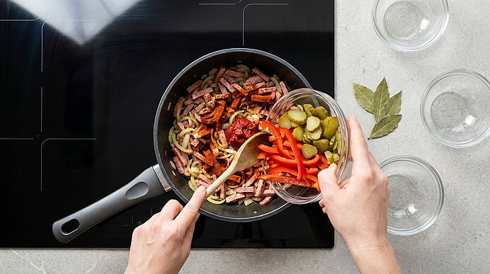 Hände geben geschnittene Gewürzgurken und rote Paprika in eine Pfanne mit angebratenen Zwiebeln und Fleischstreifen; mit einem Holzlöffel wird gerührt.